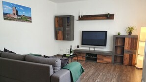 Een moderne woonkamer met een grijze bank, een houten entertainmentkast, een flatscreen televisie en een schilderij van een historisch pand.