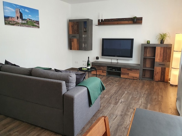 Ein modernes Wohnzimmer mit grauem Sofa, einer Holz-Unterhaltungsecke, einem Flachbildfernseher und einem Gemälde eines historischen Gebäudes.