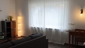 Een woonkamer met een bank, een tafel en een stoel. Er staat een lamp op de tafel en een plant aan de linkerkant.
