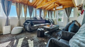 Ein geräumiges Wohnzimmer mit hoher Holzbalkendecke, großen Fenstern, einer Sofagarnitur, einem Couchtisch mit Früchten und Gläsern sowie einem weißen Heizkörper.