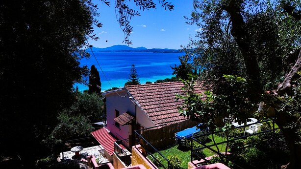 Ein Haus mit rotem Ziegeldach, einer kleinen Terrasse mit Tisch und Stühlen und Blick auf ein blaues Gewässer sowie Berge in der Ferne.