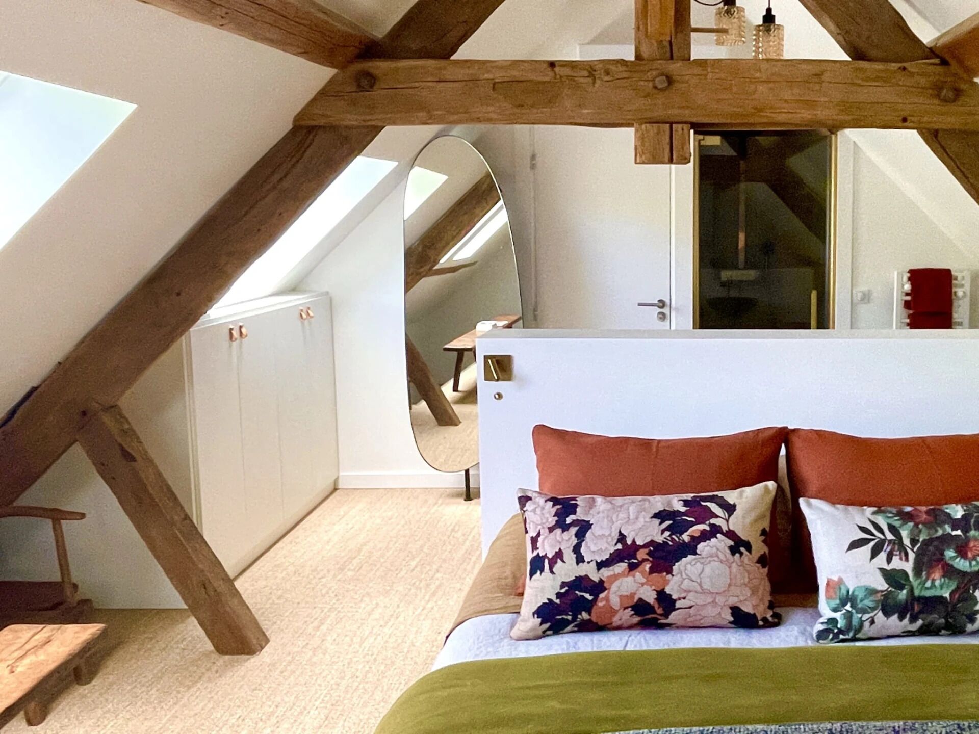 Une chambre avec un plafond en pente, un lit agrémenté d’oreillers à motifs floraux, un banc en bois et une vue sur un escalier.