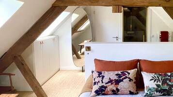 Une chambre avec un plafond en pente, un lit agrémenté d’oreillers à motifs floraux, un banc en bois et une vue sur un escalier.