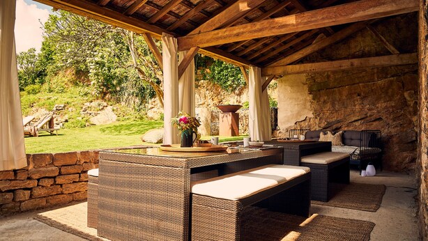An outdoor patio with wicker furniture, a stone wall, and a view of a garden. - Villa Figue Blanche (Montagny-lès-Buxy)