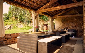 Un patio exterior con mobiliario de mimbre, una pared de piedra y vistas a un jardín.
