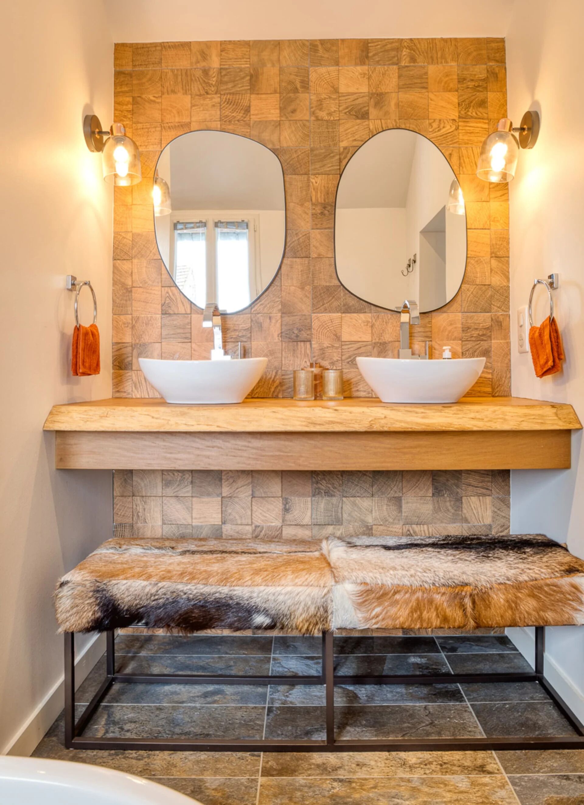 Un baño con dos lavamanos tipo pedestal, un mueble de tocador de madera, un espejo grande y un banco de piel.