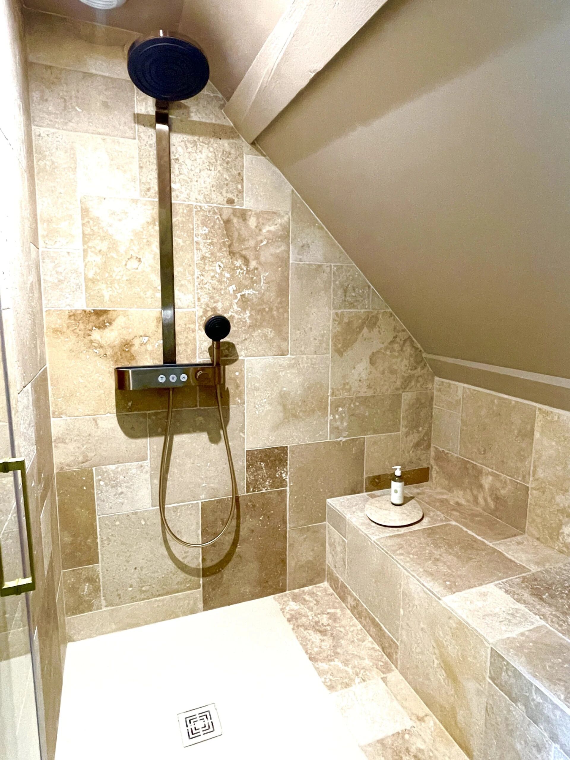 A shower area with a handheld showerhead, a wall-mounted control panel, and a soap dispenser on a small shelf.