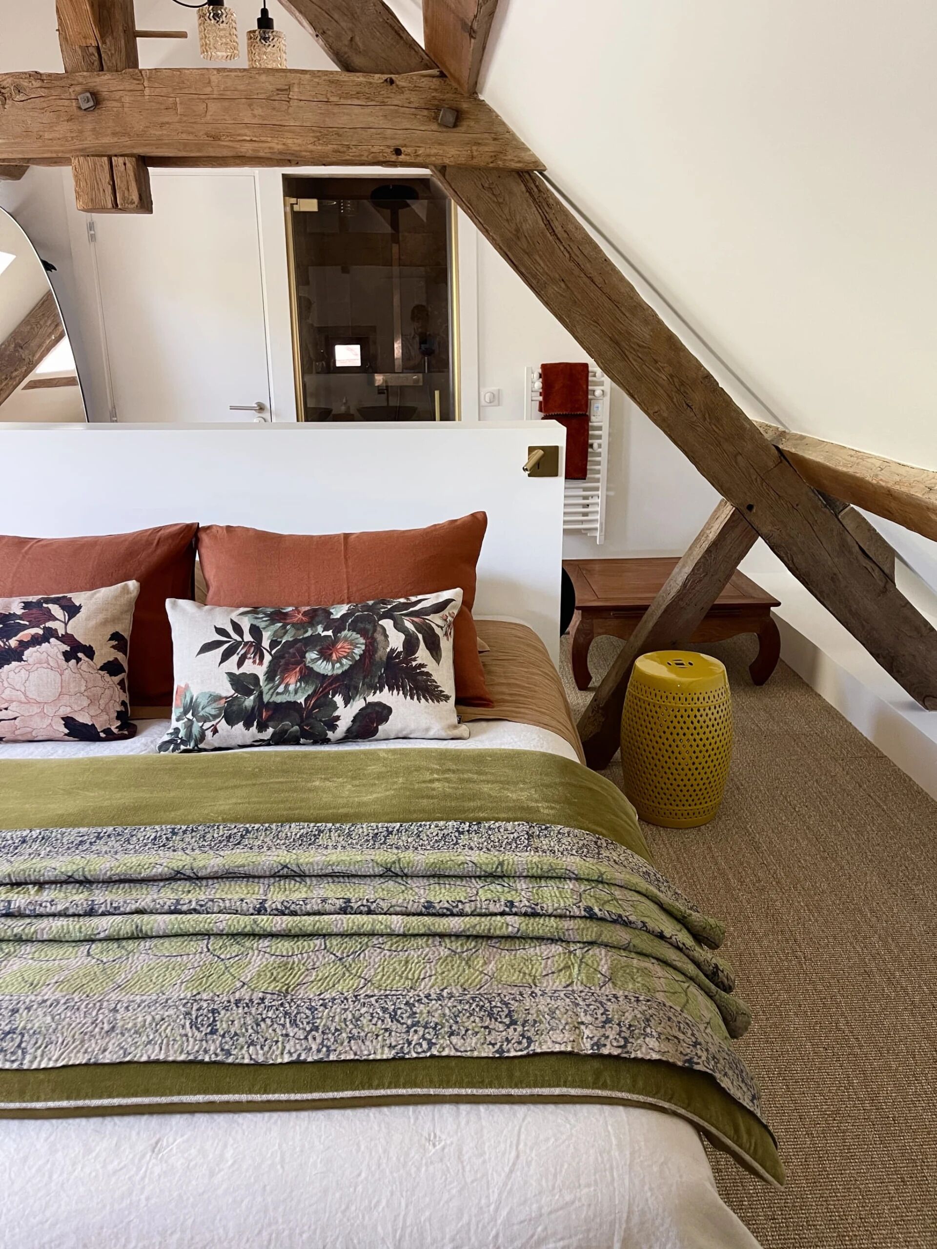 Un dormitorio con techo inclinado, una cama con almohadas estampadas y de colores lisos, una manta verde y un reposapiés amarillo.