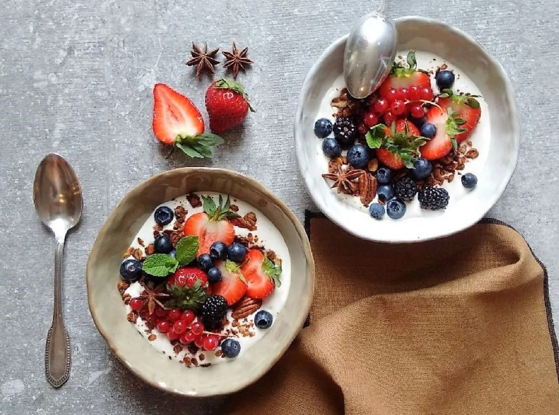 Two bowls of yogurt with mixed berries and granola on a textured surface.