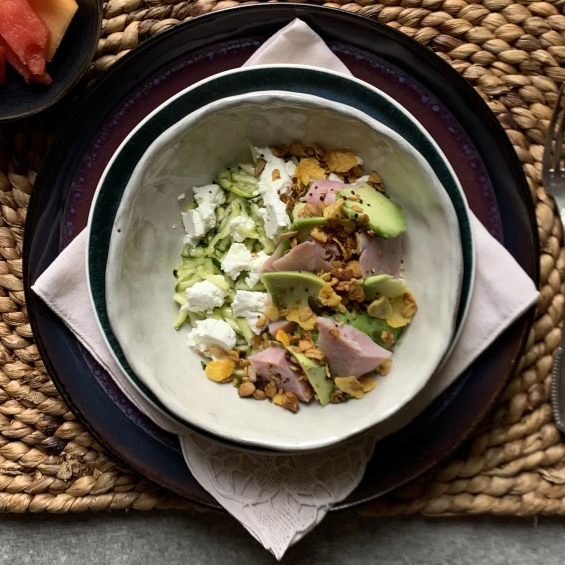 A bowl of salad with cucumbers, feta cheese, avocado, and nuts on a plate with a woven placemat.