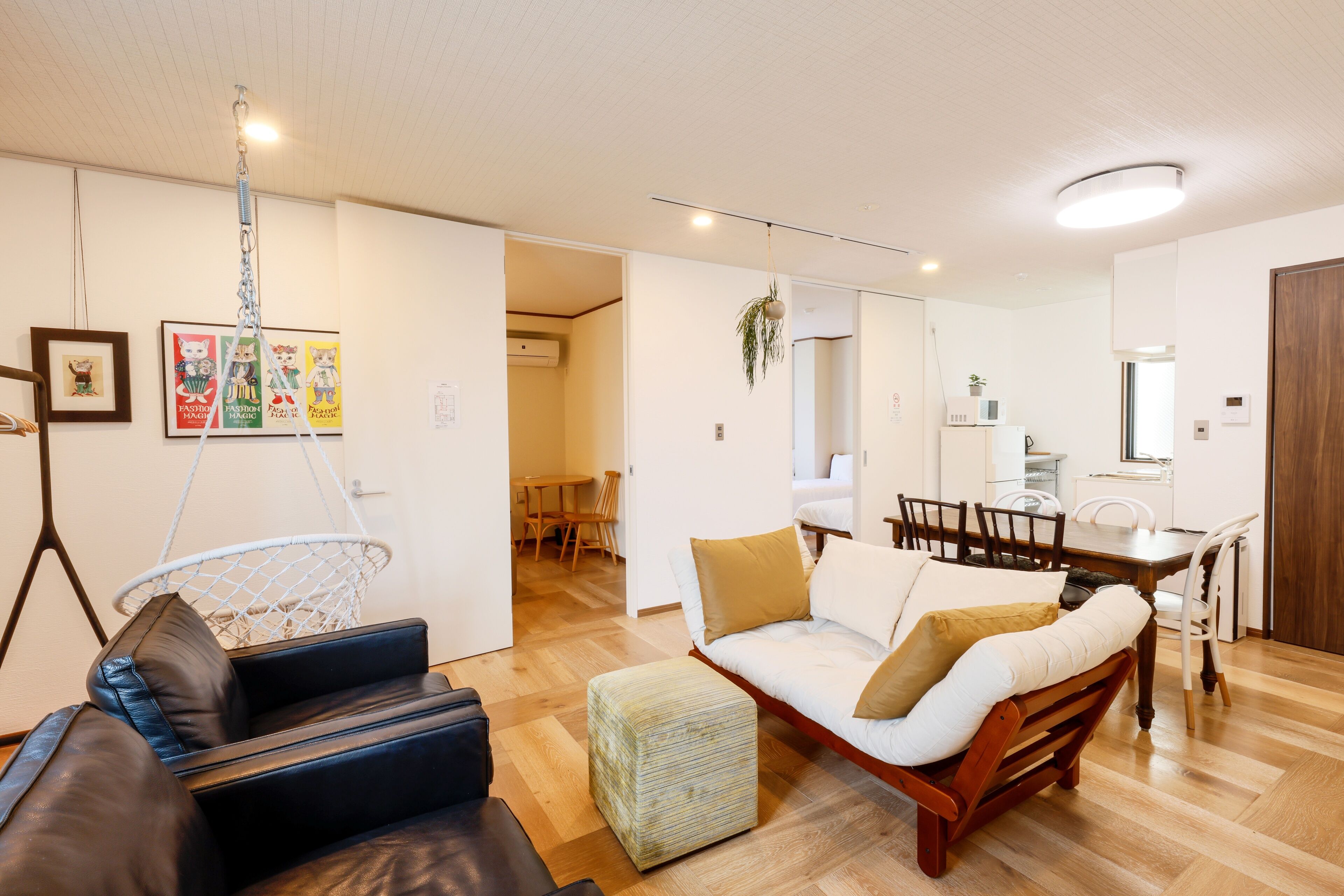 A modern living room with a sofa, ottoman, and dining table. There is a hanging plant, a ceiling light, and a door leading to another room.