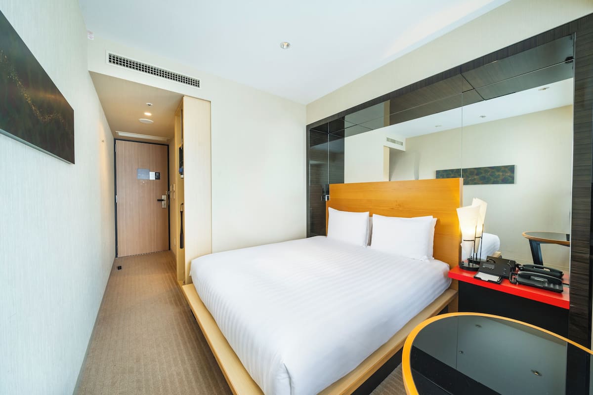 Hotellihuone, jossa on suuri sänky, yöpöytä lampun kanssa, peili ja punaisella värillä korostettu seinä.
