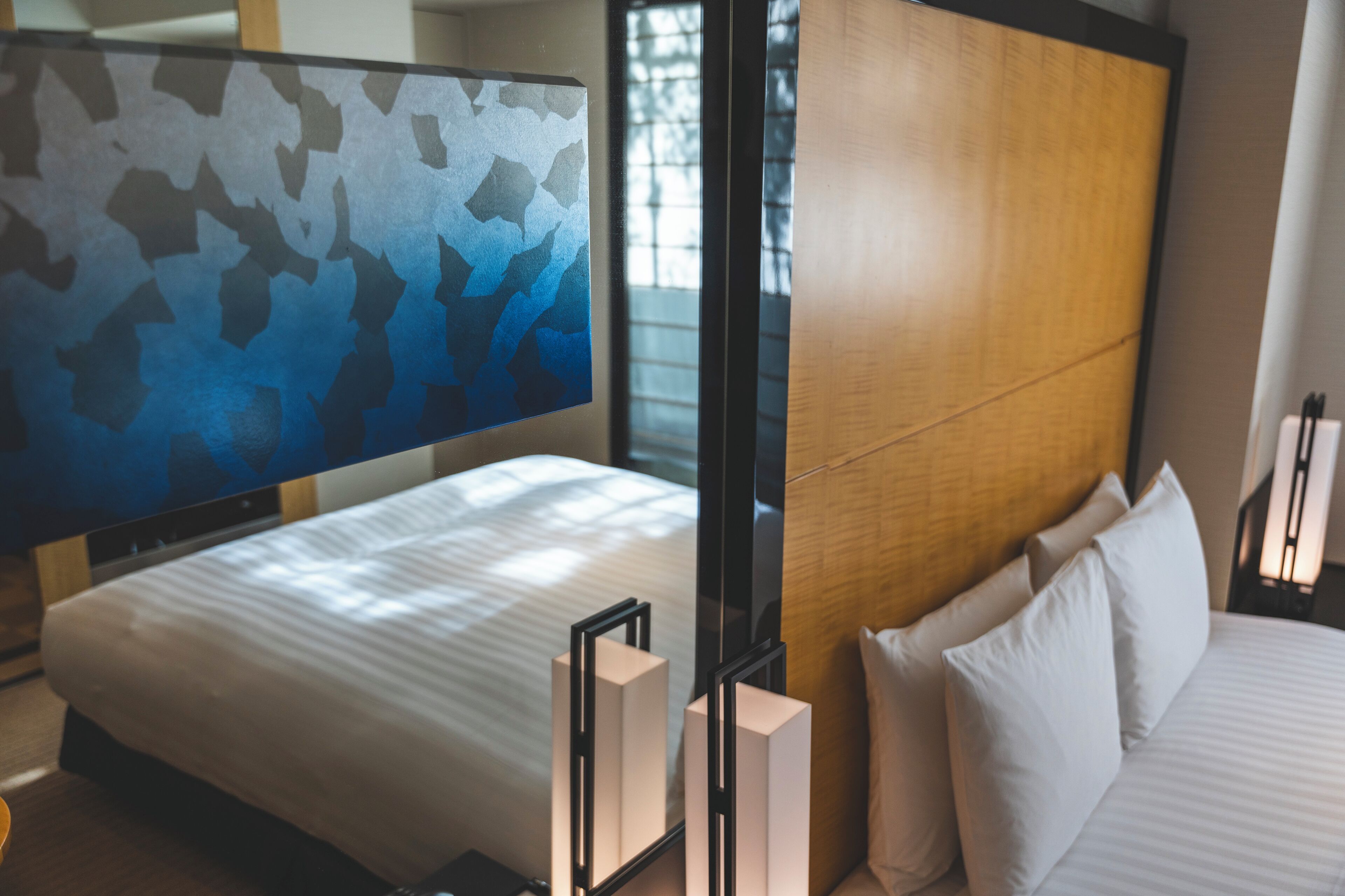 A modern hotel room with a large bed, a headboard with a wooden panel, a large abstract painting on the wall, and a bedside table with a lamp.