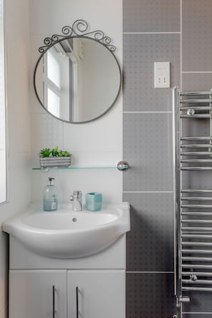 Un baño con un lavabo blanco, un espejo con marco decorativo, una estantería de vidrio con plantas, un perchero para toallas y un inodoro.