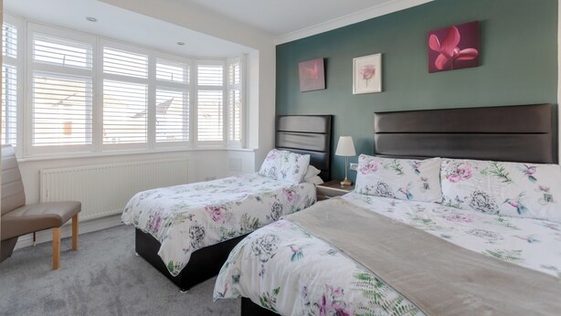 A bedroom with two beds, a window with blinds, a chair, and artwork on the wall. - Room-apple House Wembley - Family Room w (Edgware)