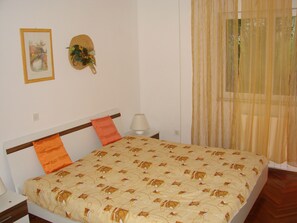 Une chambre à coucher avec un lit, une table de chevet, une lampe, une fenêtre avec des rideaux légers et un tableau encadré au mur.