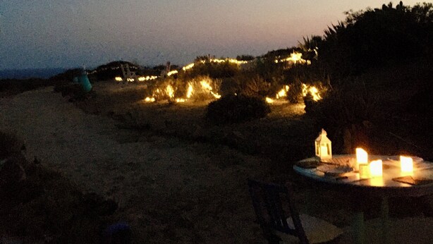 Escena de playa al anochecer con una mesa y velas, rodeada de arena y vegetación escasa.