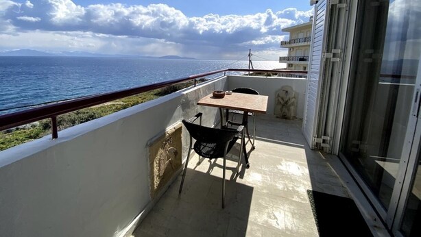 En balkon med et bord og stole, udsigt over vand og en fjerntliggende kystlinje.