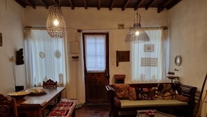 Una sala de estar acogedora con piso de madera, un sofá, una mesa de centro y una mesa de comedor con sillas. Hay dos lámparas colgantes y una ventana con cortinas.