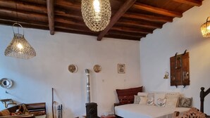 Una habitación rústica con una mesa de madera, lámparas colgantes, una cama con almohadas, una pequeña estufa y una silla de madera.