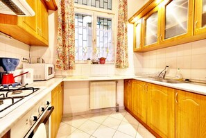 A kitchen with wooden cabinets, a gas stove, a microwave, and a window with floral curtains. - Vibrant 3 Bedroom Apartment In The Pulsing Heart Of Budapest (Budapest)