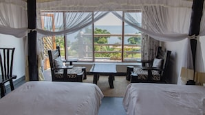 Una habitación con dos camas, una mesa y sillas, y un amplio ventanal que ofrece vistas al mar y al verdor.