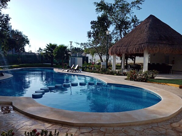 A swimming pool with lounge chairs, a thatched-roof structure, and surrounding greenery.