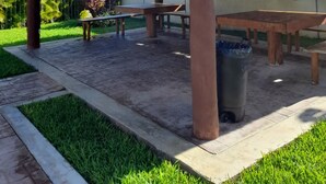 A covered outdoor seating area with a thatched roof, concrete flooring, and a trash bin.