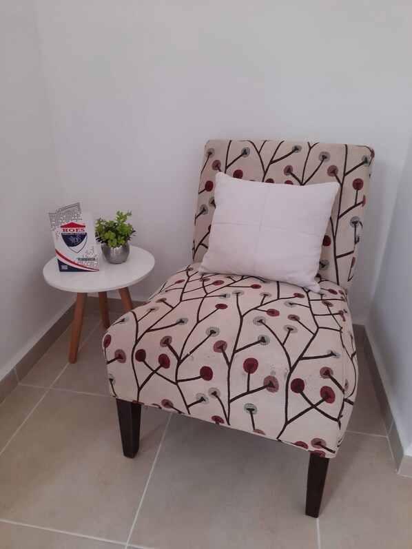 Una silla con estampado floral, un cojín blanco, una pequeña planta en maceta y una señal con texto sobre una mesa blanca.