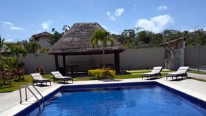 A rectangular swimming pool with a clear blue surface, surrounded by a concrete deck. There are lounge chairs and a thatched-roof structure in the background.