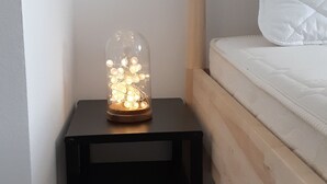 Un lit avec une tête de lit en bois et une literie blanche, une petite table de chevet noire sur laquelle se trouve un bocal en verre contenant des guirlandes lumineuses, et un mur blanc uni.
