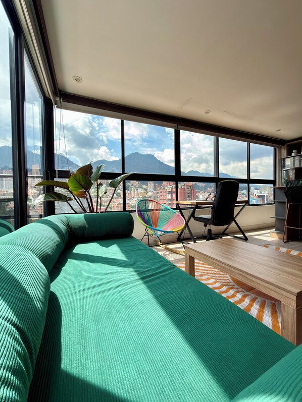 A modern living room with a large window, a green sofa, a colorful chair, and a wooden table.