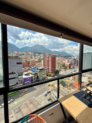 A cityscape view from a high-rise window, featuring a mix of modern and older buildings, a busy street with vehicles, and a mountain range in the background.