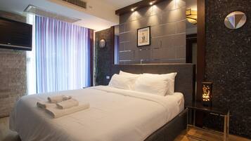 A modern hotel room with a large bed, a flat-screen TV, a nightstand with a lamp, and a view of the city through the window.