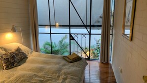 Une chambre avec une grande fenêtre, un lit, une lampe et une vue sur la verdure environnante.