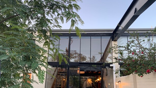 Un bâtiment moderne doté de grandes fenêtres en verre, d’un escalier en bois menant à un espace intérieur et d’un coin salon extérieur.