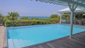 A clear blue pool with steps leading into it, surrounded by a wooden deck and a covered area with white lattice detailing. - VILLA HERMES "dieu des voyageurs " (SAINTE ANNE)