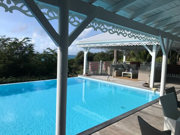 Una piscina azul cristalina con un pabellón blanco, rodeada de exuberante vegetación y vista al océano.