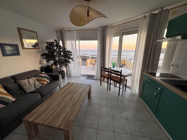 A living room with a sofa, a coffee table, a dining table with chairs, a large potted plant, and a view of the sea through the windows.