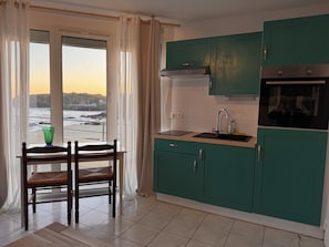 A kitchen with teal cabinets, a window with curtains, a dining table with chairs, and a view of the sea.
