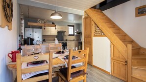 A dining area with wooden chairs and a table set for two, a kitchen in the background, and a wooden staircase.