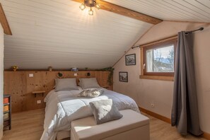 Une chambre avec un plafond en pente, une tête de lit en bois, un lit avec des oreillers, une table de chevet, une fenêtre avec des rideaux et un tableau accroché au mur.