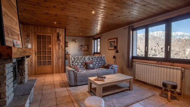 A cozy living room with a stone fireplace, a sofa with patterned cushions, a wooden coffee table, and a view of mountains through large windows.