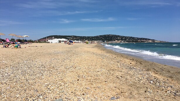 A pebbled beach with umbrellas and beach chairs, a hill in the background, and a clear sky.