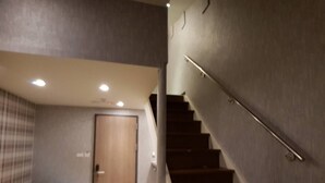 Un escalier avec une rampe, une porte en bois et un canapé jaune dans une pièce dont le mur est texturé et qui est éclairée par des luminaires au plafond.
