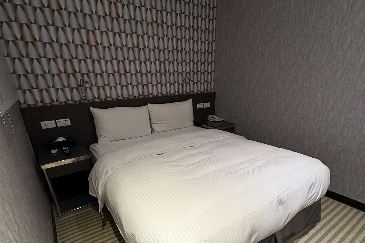 Una habitació d'hotel amb un llit, una vetustat, un telèfon i una paret amb un dibuix geomètric.
