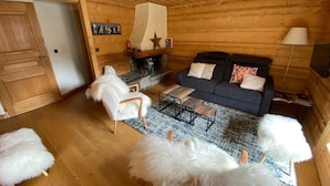 L’intérieur d’un chalet confortable, avec des murs en rondins de bois, une cheminée en pierre, un canapé sombre, des fauteuils moelleux blancs et un plancher en bois.