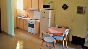A compact kitchen with a stove, microwave, and refrigerator. There is a dining table with chairs and a clock on the wall. - Alkioni City Apartments (Chania)