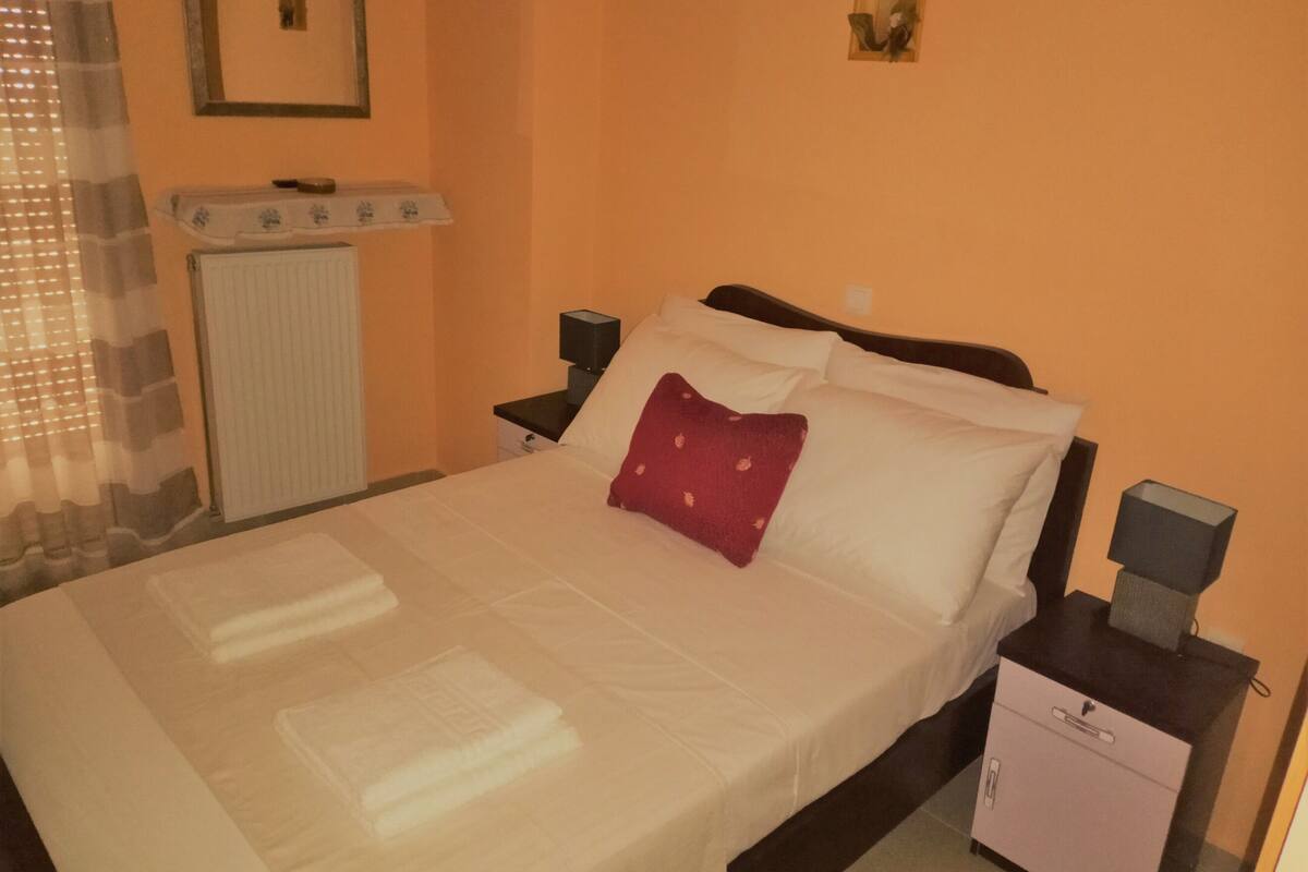 A neatly made bed with white linens and a red pillow, a nightstand with a lamp, a small cabinet, and a radiator on the wall.