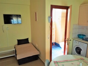 A small, compact room with a bed, a television mounted on the wall, a washing machine, and a sink. - Alkioni City Apartments (Chania)